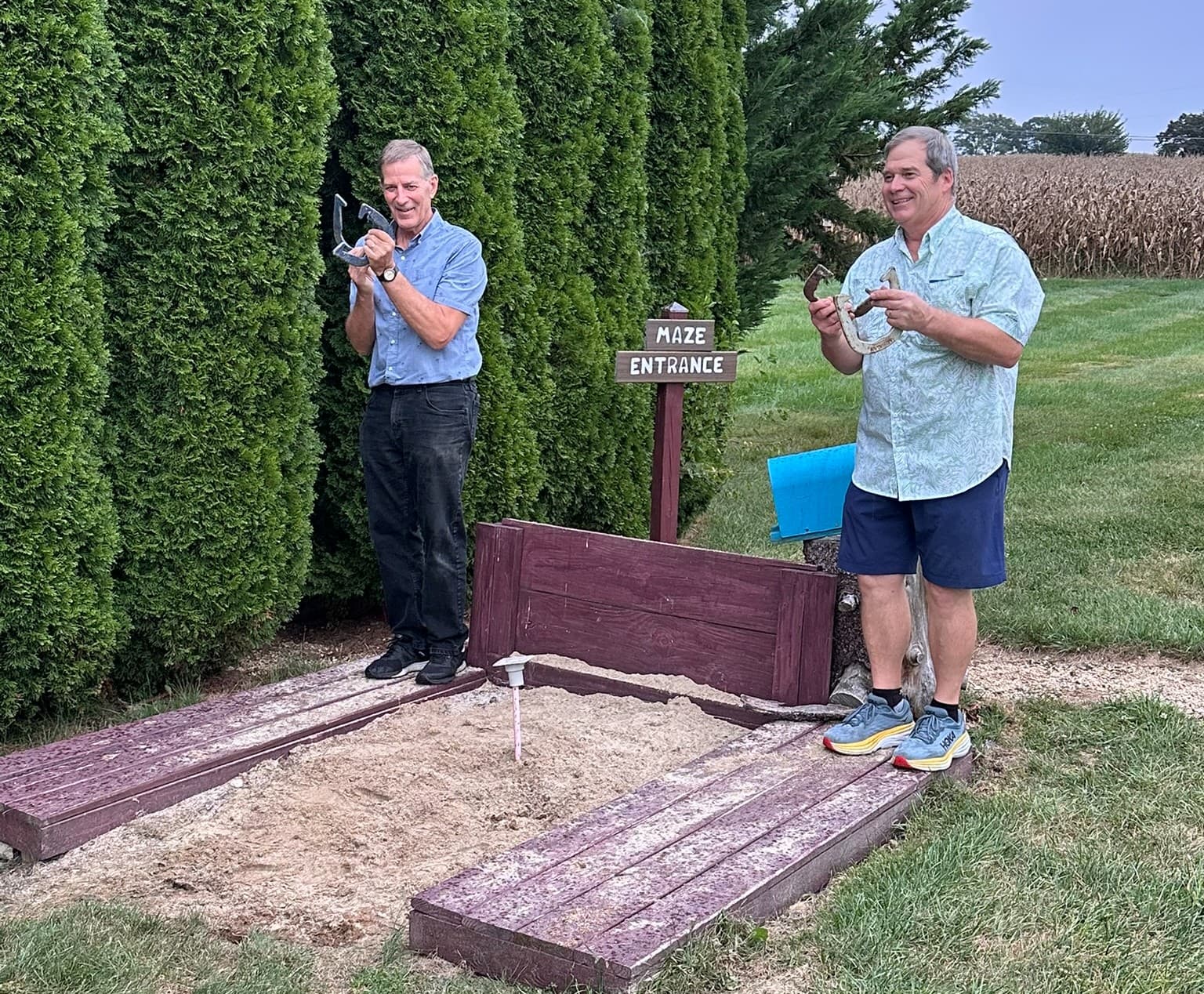 Two men stand at a maze entrance, holding tools and smiling, surrounded by greenery and a field in the background.