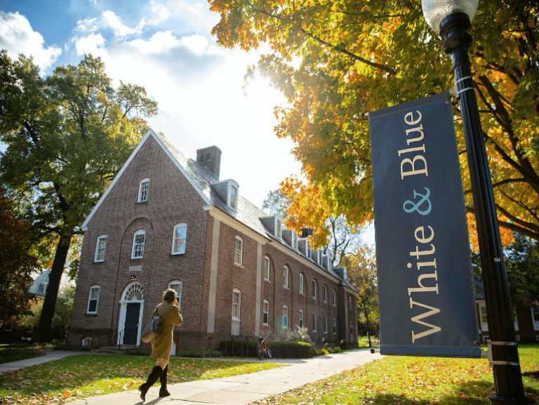 A person walks past a brick building adorned with a "White & Blue" banner, surrounded by autumn foliage.