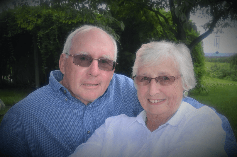 An elderly couple smiles together outdoors, surrounded by greenery.