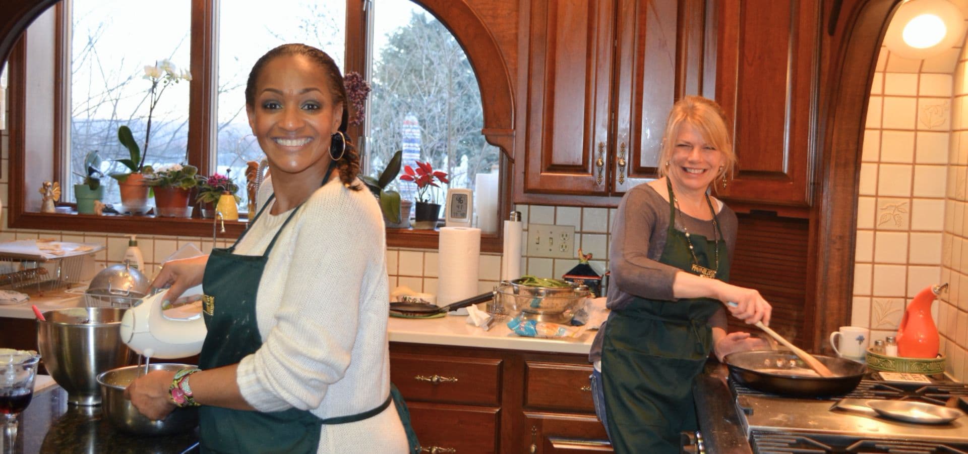 Two women in aprons smile while cooking together in a warm kitchen filled with plants and cooking utensils.