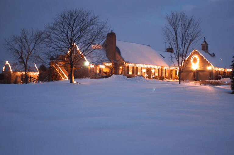 A snow-covered house adorned with warm holiday lights under a twilight sky.