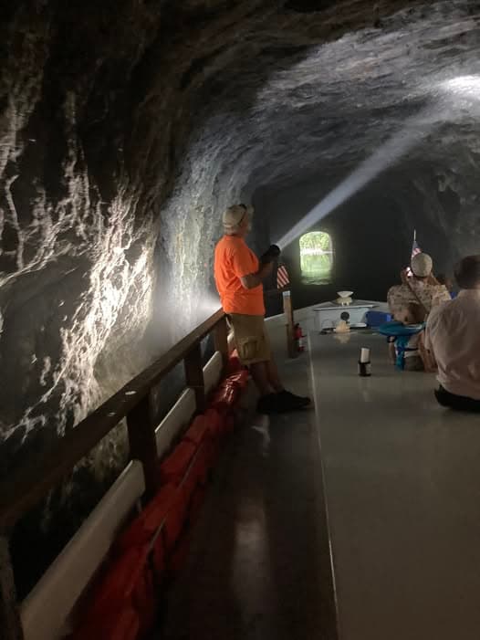 A guide shines a flashlight inside a rocky tunnel while visitors sit at a table on a boat.