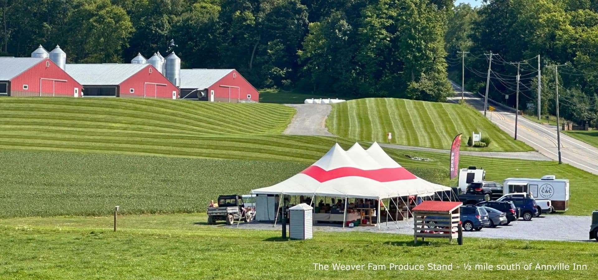 A vibrant farm scene featuring a produce stand under a tent, surrounded by green fields and red barns.