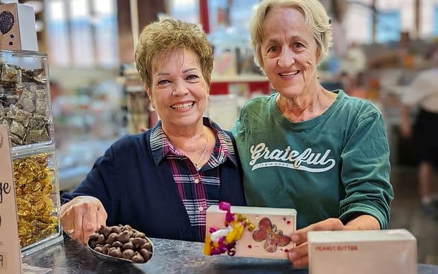 Two smiling women stand behind a counter, one holding a tray of chocolates and the other showing a decorated box.