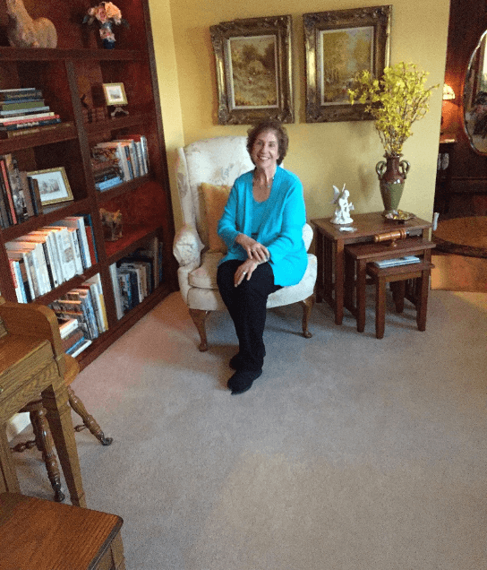 A smiling woman in a blue sweater sits comfortably in an armchair, surrounded by books and decorative items in a cozy living room.