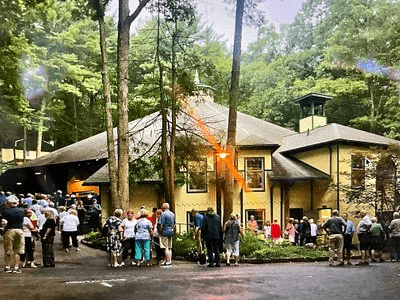 A crowd gathers outside a large, yellow building surrounded by trees.