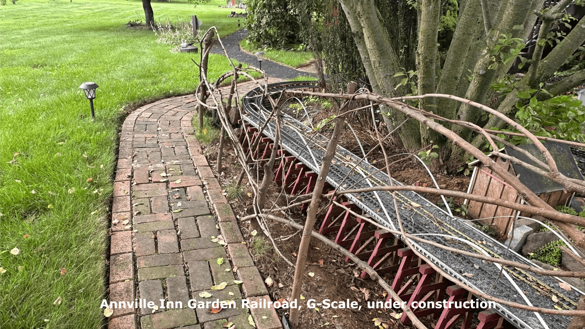 A winding brick path beside a G-Scale garden railroad under construction, surrounded by grass and trees.