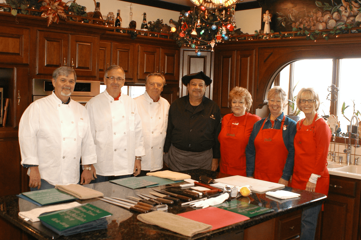 A group of seven people, including chefs in white coats and others in red aprons, stand together in a festive kitchen.