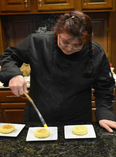 A person in a black chef's coat uses a kitchen torch on a dessert in a kitchen.