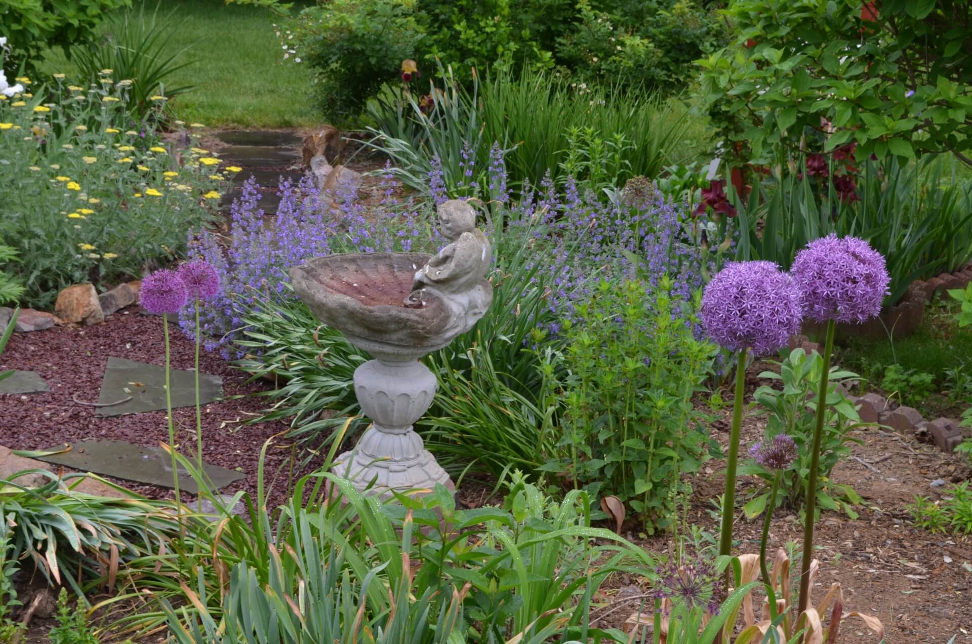 A serene garden featuring purple flowers and a stone birdbath with a figure.