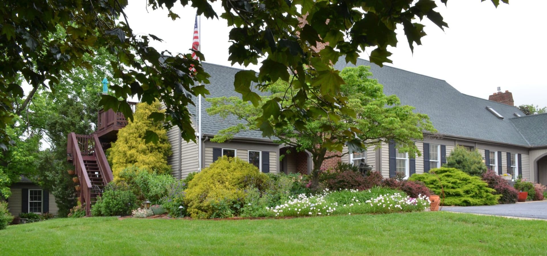 A well-maintained house surrounded by lush greenery and flower beds, with an American flag visible.