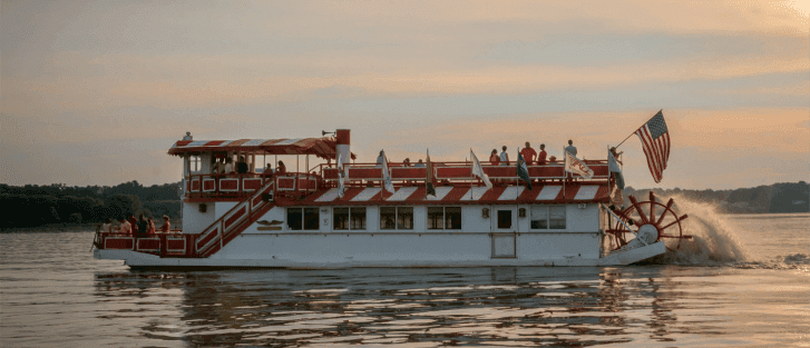 A paddleboat with red and white accents cruises on calm waters at sunset.