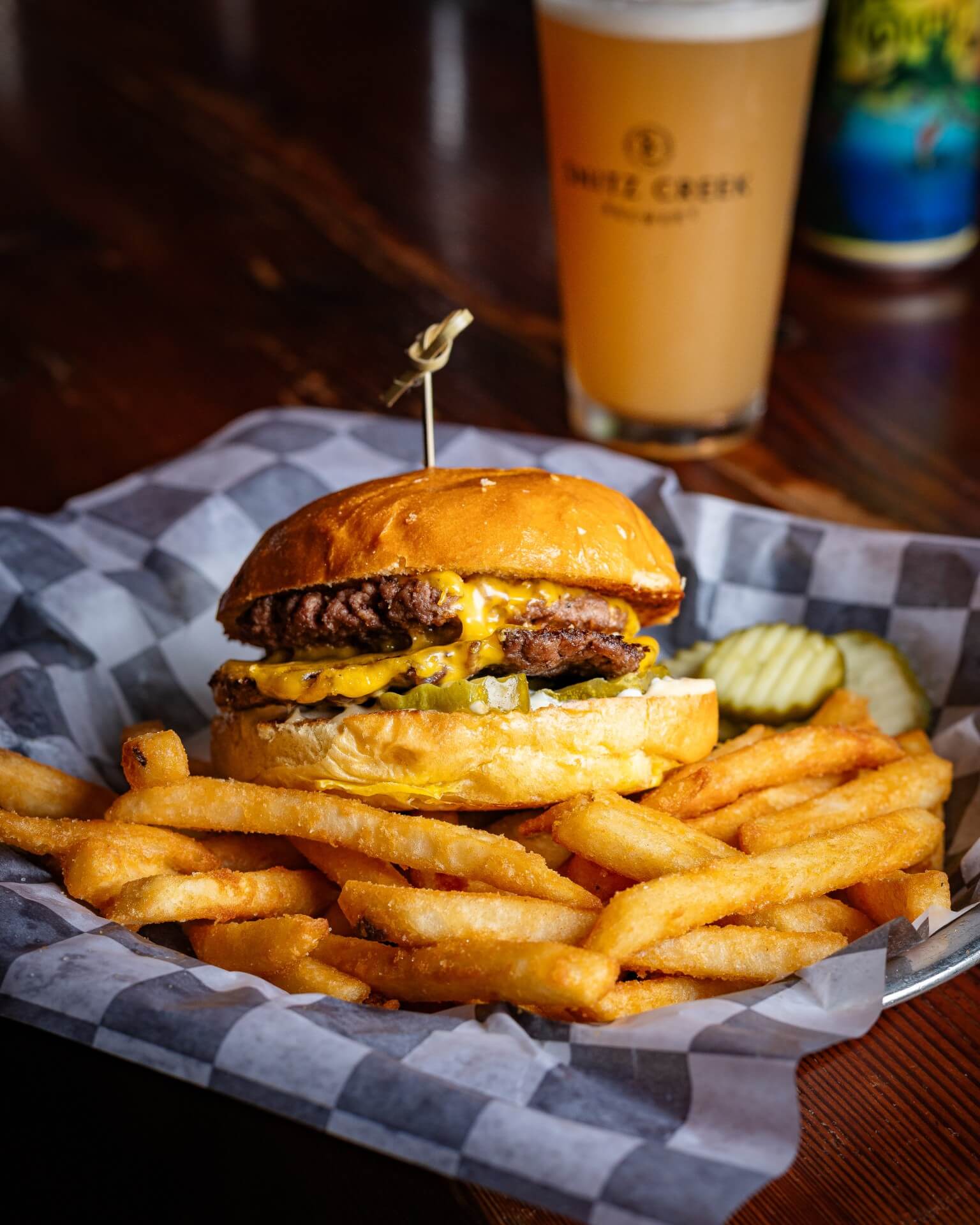 A cheeseburger with pickles sits on a plate alongside crispy French fries and a glass of beer in the background.