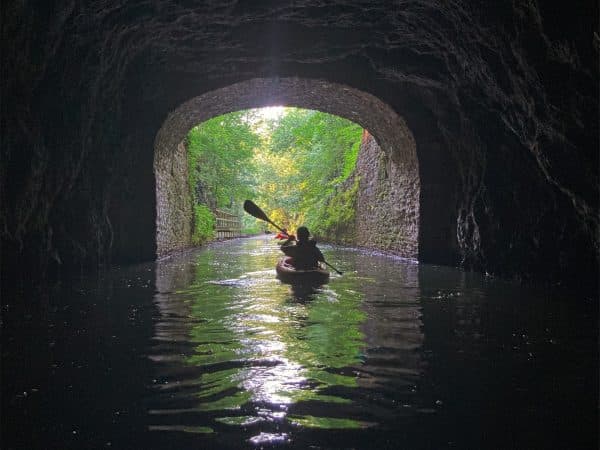 A kayaker paddles through a dark tunnel, emerging into a bright, green landscape.