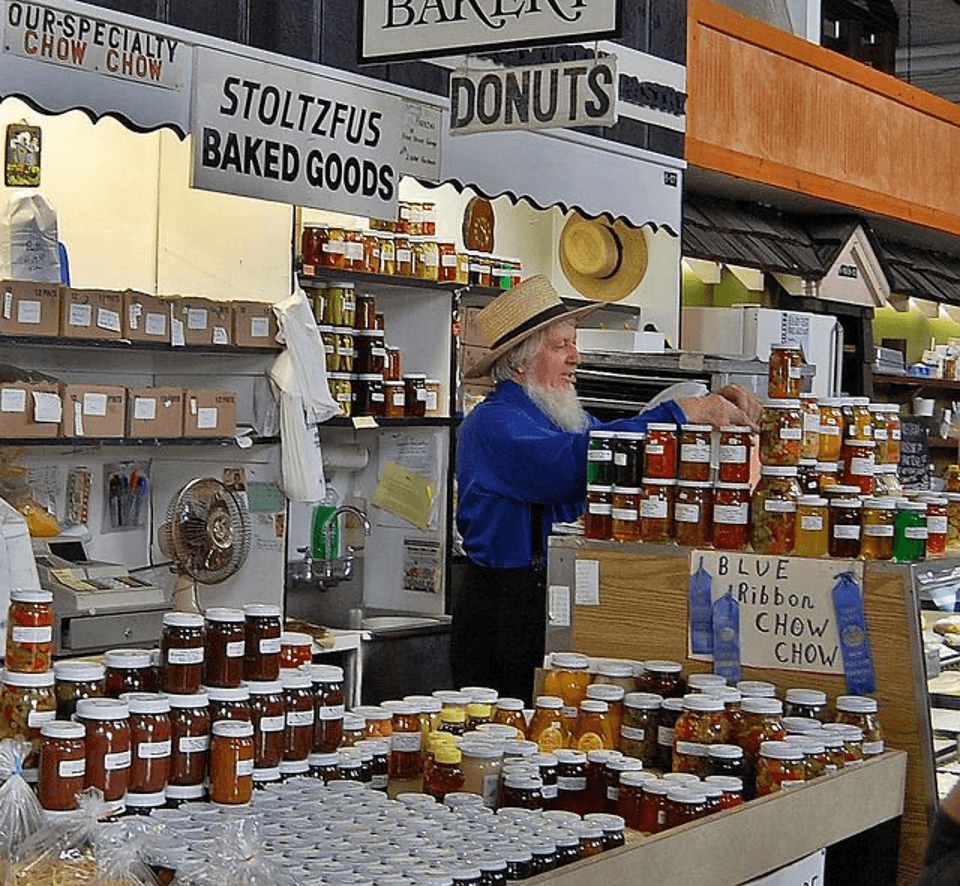 A vendor in a straw hat arranges jars of preserves at a market stall labeled "Stoltzfus Baked Goods."