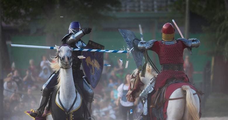 Two armored knights on horseback engage in a jousting match, with spectators in the background.