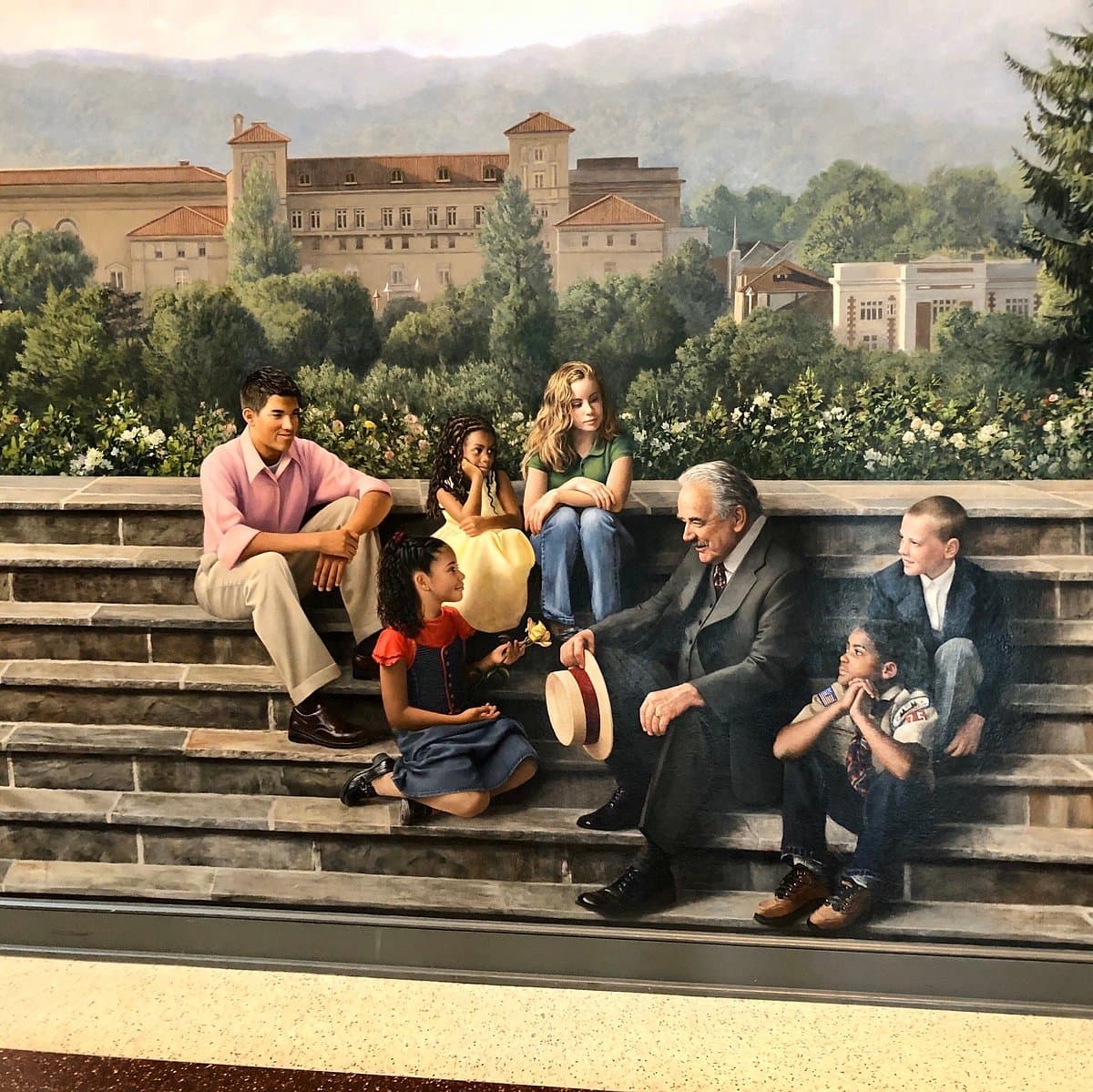 A mural depicting a group of diverse children engaged in conversation with an older man on a staircase, set against a backdrop of buildings and trees.