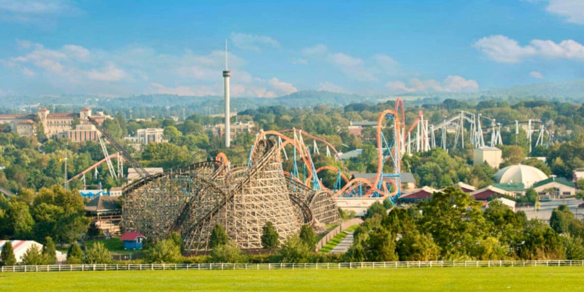 A panoramic view of a theme park featuring a wooden roller coaster and various rides against a blue sky.