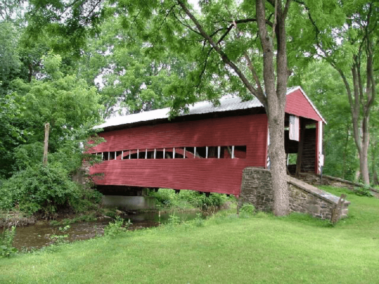 A red covered bridge spans a small stream, surrounded by lush greenery.