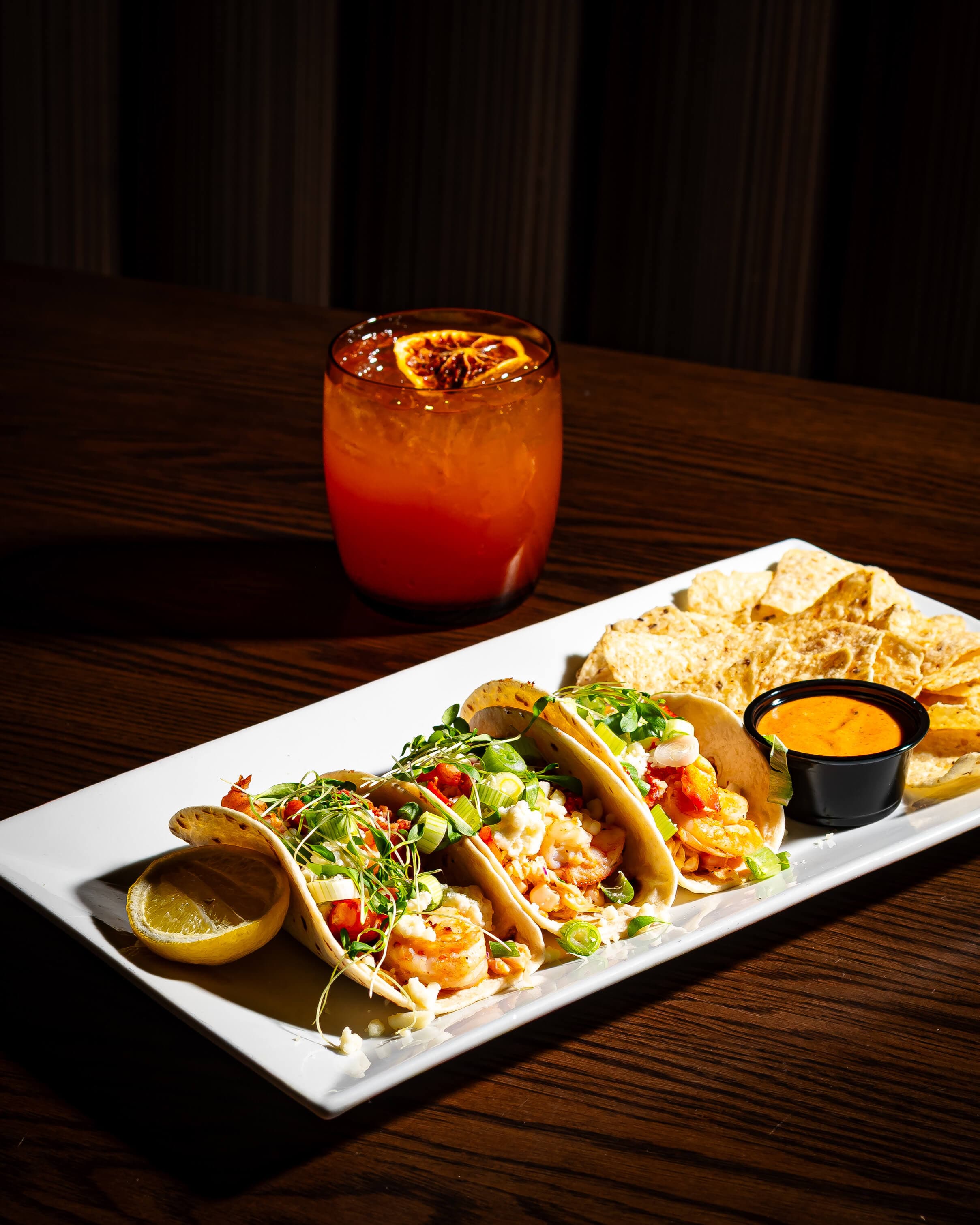 A plate of shrimp tacos garnished with herbs and vegetables, accompanied by tortilla chips and a cocktail.