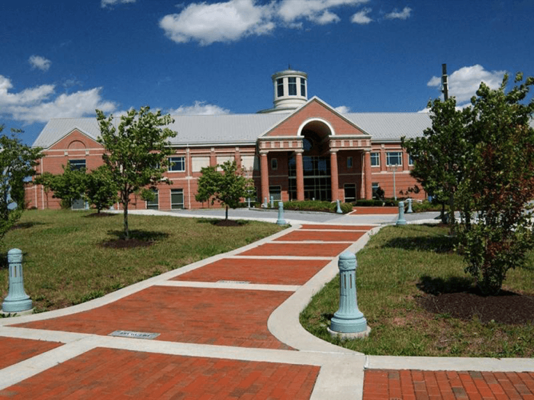 A brick pathway leads to a modern building with a prominent cupola, surrounded by greenery and blue skies.