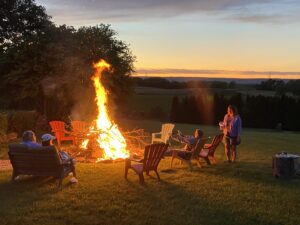 A group of people sitting around a bonfire near sunset with Pennsylvania countryside in the background.