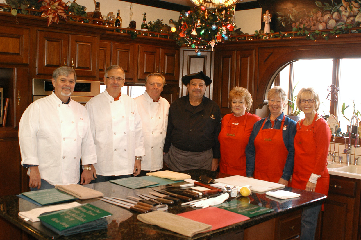 Chef in black chef's outfit with six students he is teaching. The men are wearing white cooking jackets and the women red aprons. Acclaimed Chef Cooking Class, Annville Inn.