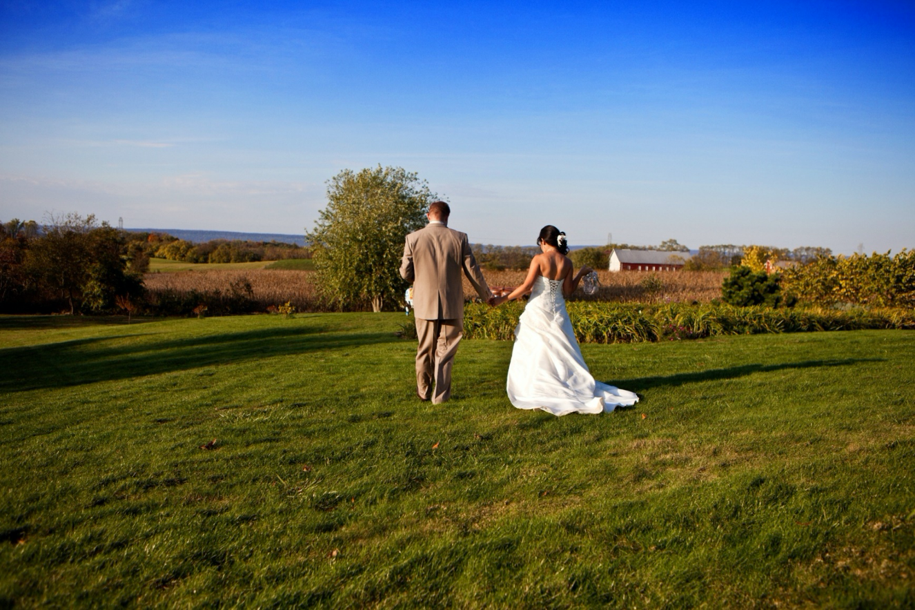 Bride and Groom walking through lawn toward old farmstead in background. Further back, the Blue Mountains and the blue sky.