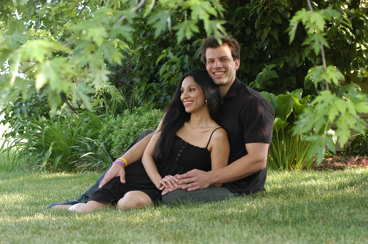 Portrait of couple outdoors: Handsome young man seated on ground with long haired young lady. Both smiling brightly.