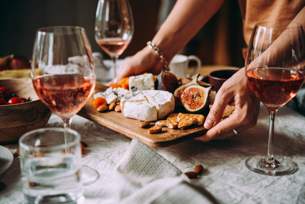 Beautiful stable setting with nice linens and one sees two glasses of wine and a glass of water.  A lady is setting a tray down among the glasses. It is laden with cheese, fruits, crackers (to accompany the wine.)
