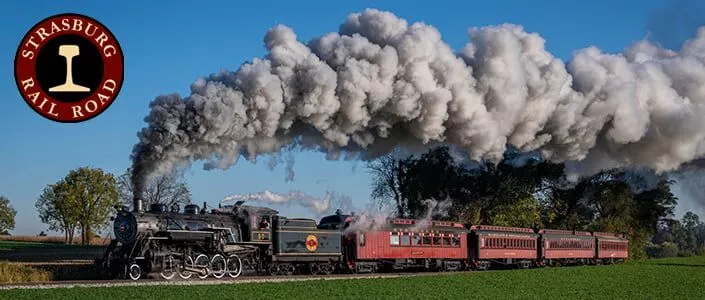 A steam locomotive pulls four passenger carriages down the tracks with a bright blue sky in the background, and a green farm field in the foreground.