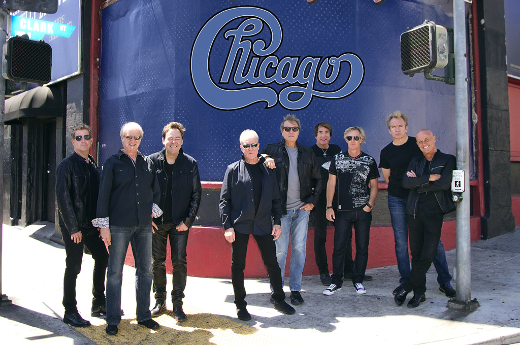 Photograph of the band, Chicago, outdoors, created within last few years, so they are older now. Large logo is above them on the building behind them.  Chicago at HersheyTheatre coming in April.