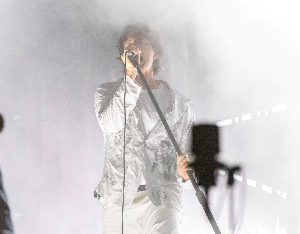 Lead singer of Third Eye Blind singing into a microphone he is holding at an angle. He is dressed in all white and the background is also in white shades. High Key. This  Hershey Pa Concert will be at Hollywood Casino.
