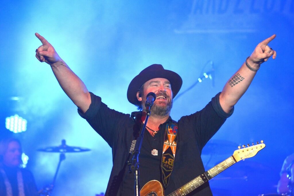 Photo of C&W singer Lee Brice, on stage during a concert, blue light bathing the stage in the background. A guitar is suspended from his neck, and he is holding both arms up int he air.  Hollywood Casino Near Hershey PA Concerts will host him this summer. He has a huge smile on his face.