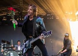 Rick Springfield holding guitar singing at a concert. In background is a drummer to his right and a guitarist to his left.  Hollywood Casino Near Hershey PA Concerts will feature him this summer.