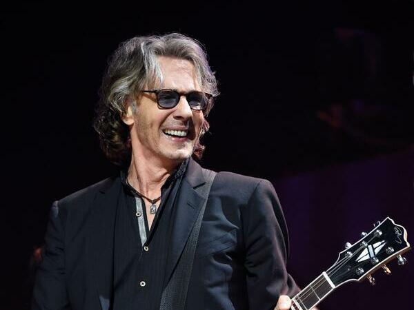 Singer Rick Springfield holding a guitar, looking to his left, smiling and singing. He is wearing sunglasses.  He has a wide open smile!  Hollywood Casino Near Hershey PA Concerts will showcase him this summer.