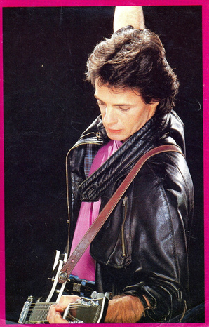 A young Rick Springfield entertains the crowd from his place on stage. He is wearing a black leather jacket, purple shirt, and is holding a guitar that is also held onto him via a strap over his right shoulder.