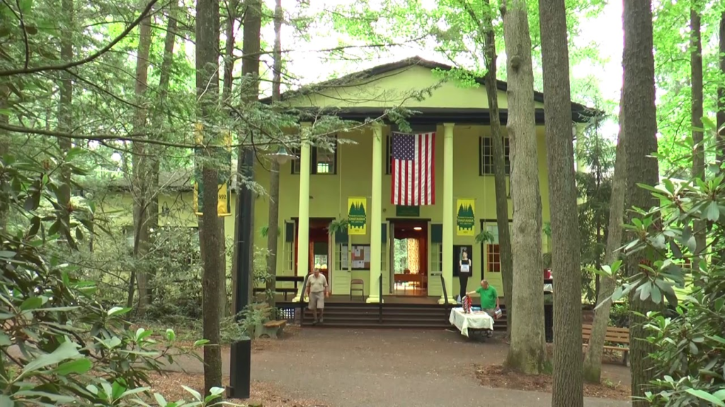 Image shows a large structure with columns in front. The building is the headquarters of the Pennsylvania Chautauqua and is called the "Hall of Philisophy."  The building's colors are a yellow-green with brown trim. It is two stories tall with an impressive pediment.  An American flag hangs down between the two center columns.