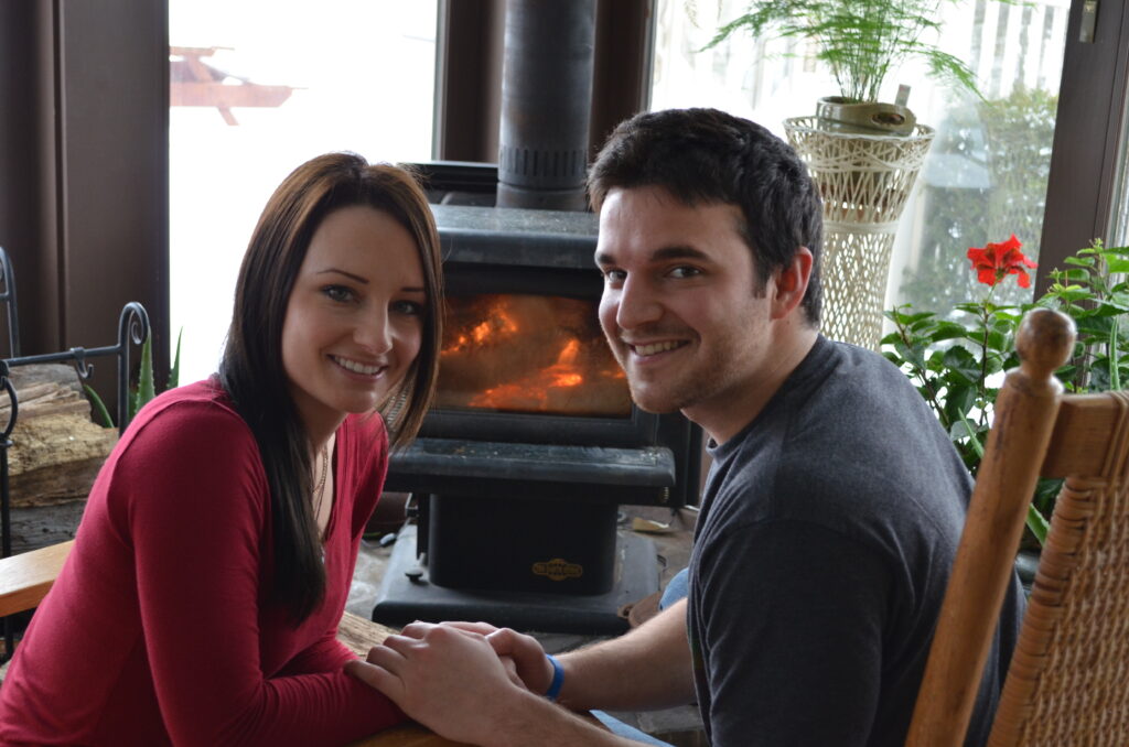 Photo of couple smiling and holding hands, in front of a fireplace